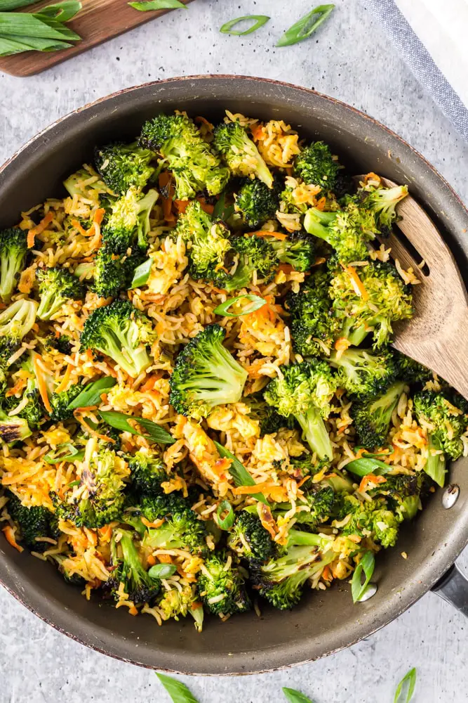 Saute pan filled with bright green broccoli florets, grated carrots, sliced green onions and fried rice