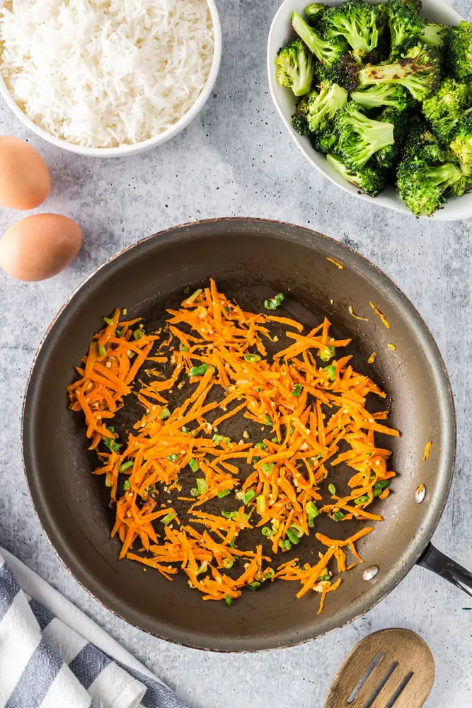 Grated carrots in a saute pan with green onions and two eggs, a bowl of cooked white rice and a bowl of green roasted broccoli on the side