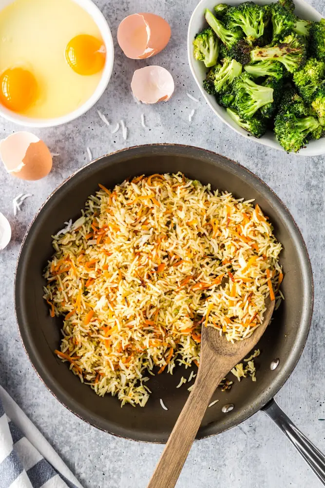 rice and and grated carrots in a saute pan being fried with two eggs cracked into a small bowl and a bowl of green roasted broccoli on the side