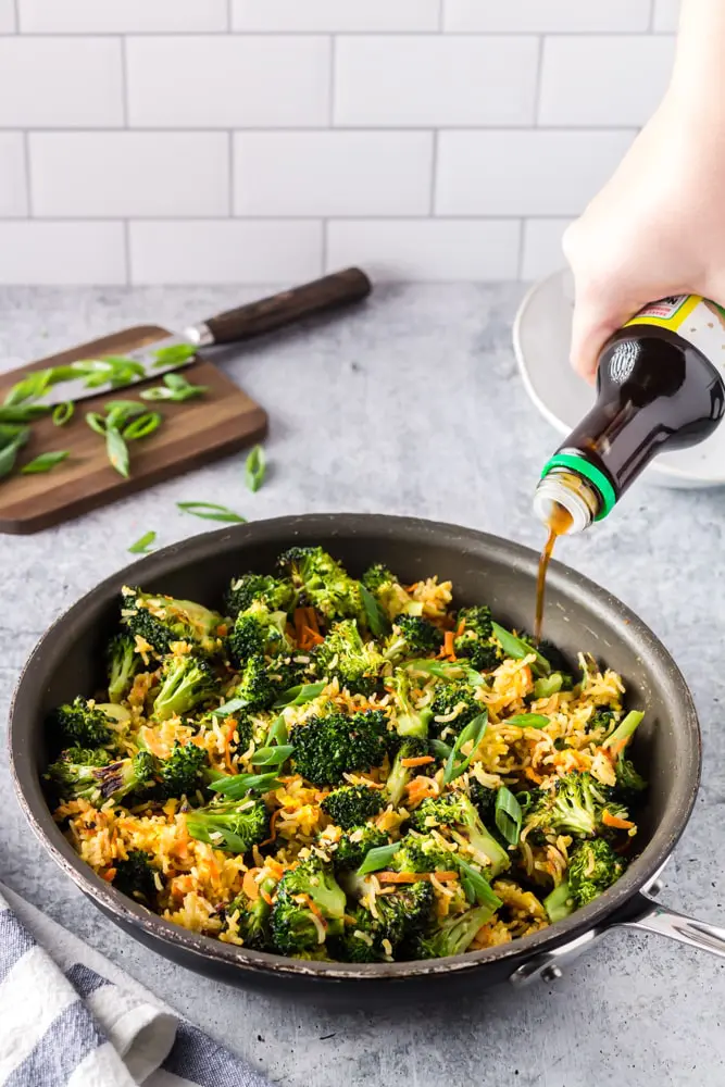 Saute pan filled with bright green broccoli florets, grated carrots, sliced green onions and fried rice with ponzu sauce being drizzled in