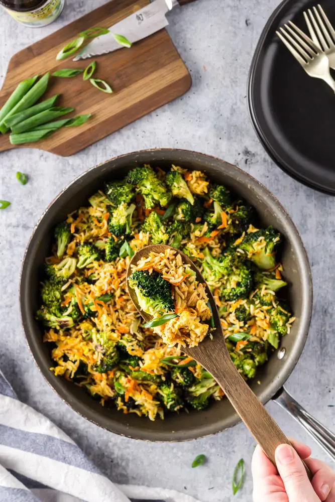 Saute pan filled with bright green broccoli florets, grated carrots, sliced green onions and fried rice with a spoon scooping out a heap