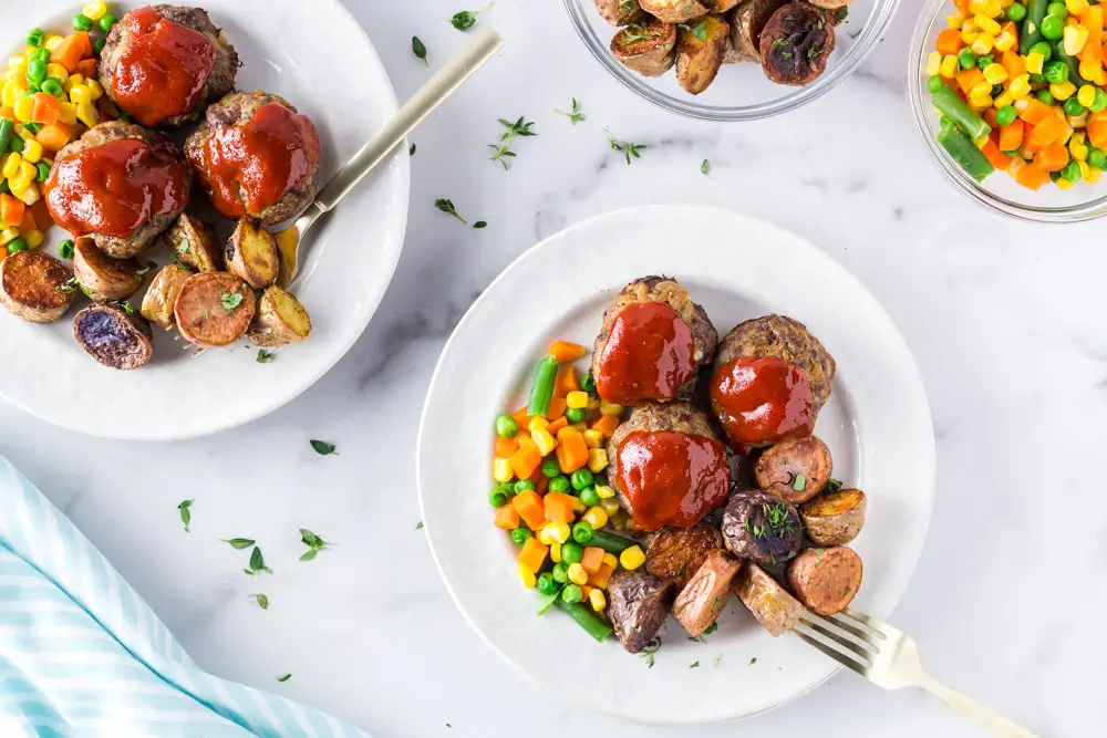 Two plates of Onion Burger Mini Meatloaf covered in a bright red sauce with carrots, pea, and green beans on the side