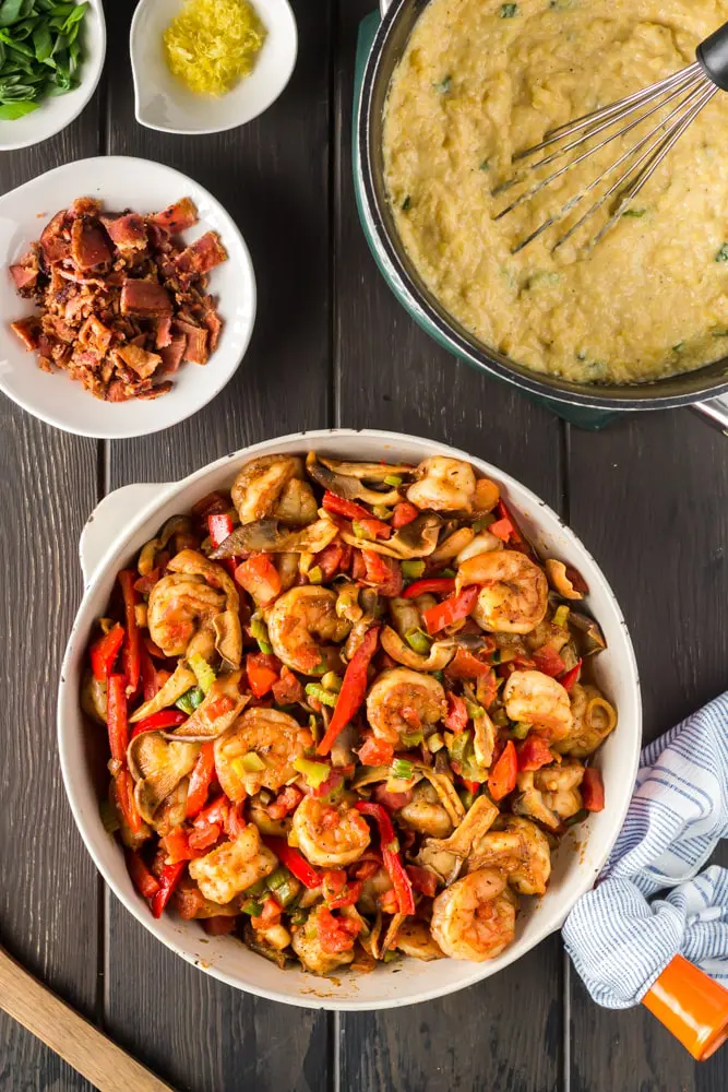 orange saute pan filled with cooked shimp, green onions, bell peppers and mushrooms with a bowl of bright yellow grits and small bowls filled with bacon, green onions and lemon zest on the side