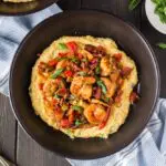 Black bowl filled with bright yellow grits, topped with shrimp smothered in a new orleans style buttery barbecue sauce and tossed with green onions, red bell peppers and mushrooms