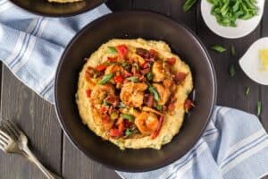 Black bowl filled with bright yellow grits, topped with shrimp smothered in a new orleans style buttery barbecue sauce and tossed with green onions, red bell peppers and mushrooms