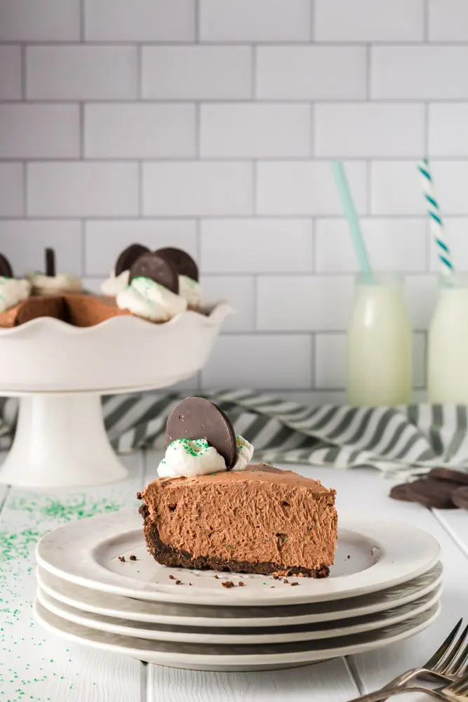 Single slice of brown thin mint chocolate cheesecake on a white plate with the whole cheesecake plate on a cake pedestal in the background