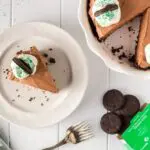 Single slice of brown thin mint chocolate cheesecake on a white plate with the whole cheesecake plate on the side missing a slice and thin mint cookies falling from the box