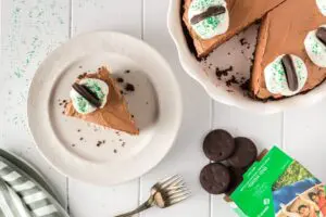 Single slice of brown thin mint chocolate cheesecake on a white plate with the whole cheesecake plate on the side missing a slice and thin mint cookies falling from the box