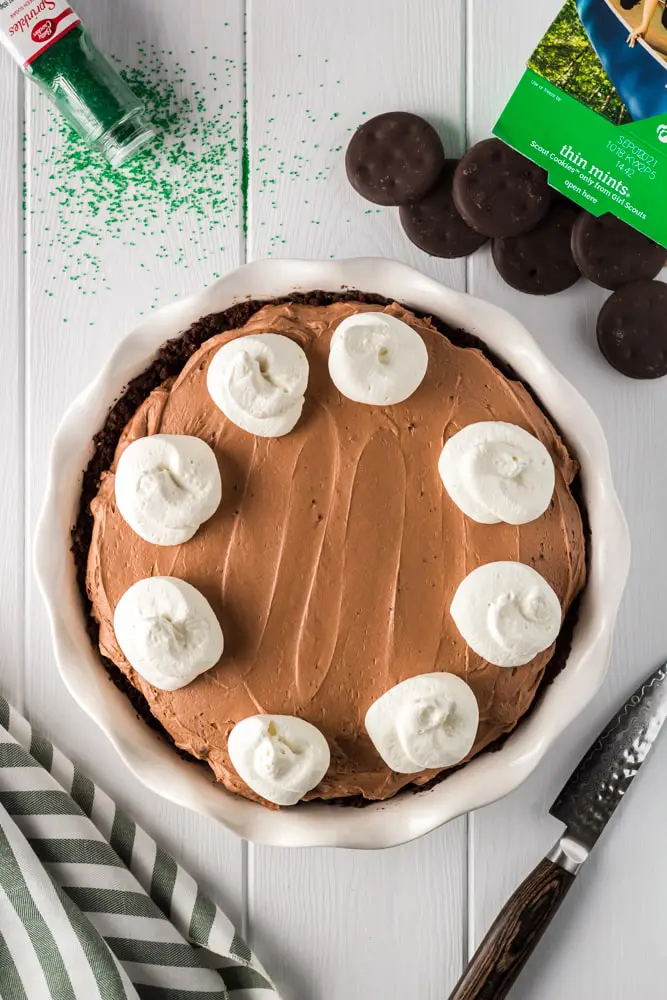 whole and unsliced brown thin mint no bake chocolate cheesecake with white dollops of whipped cream on each slice with thin mint cookies and green sprinkles on the side before adding