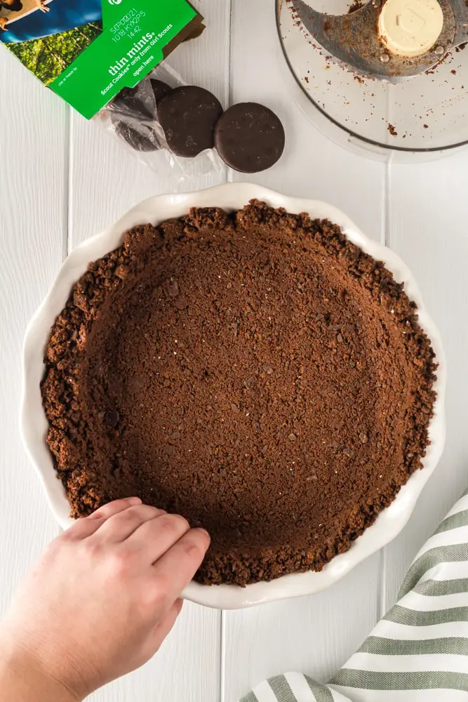 unbaked thin mint cookie crust being pressed by hand into a white pie plate