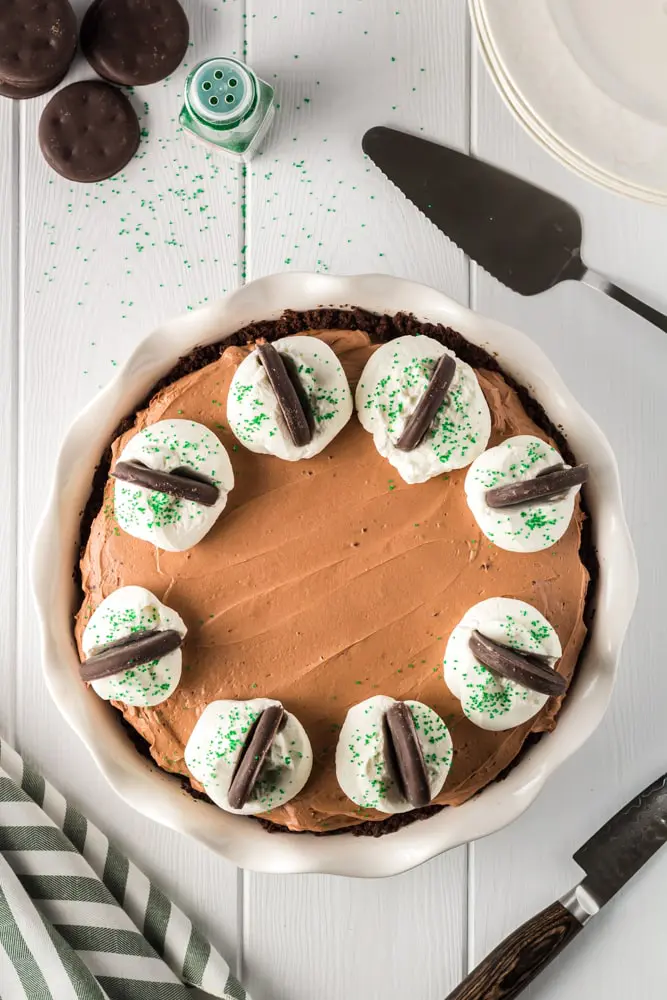 whole and unsliced brown thin mint no bake chocolate cheesecake with white dollops of whipped cream on each slice topped with bright green sprinkles and a thin mint cookies