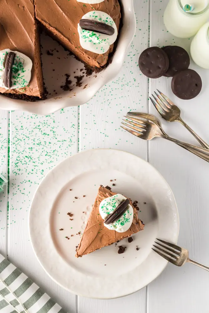 Single slice of brown thin mint chocolate cheesecake on a white plate with the whole cheesecake plate on a cake pedestal in the background