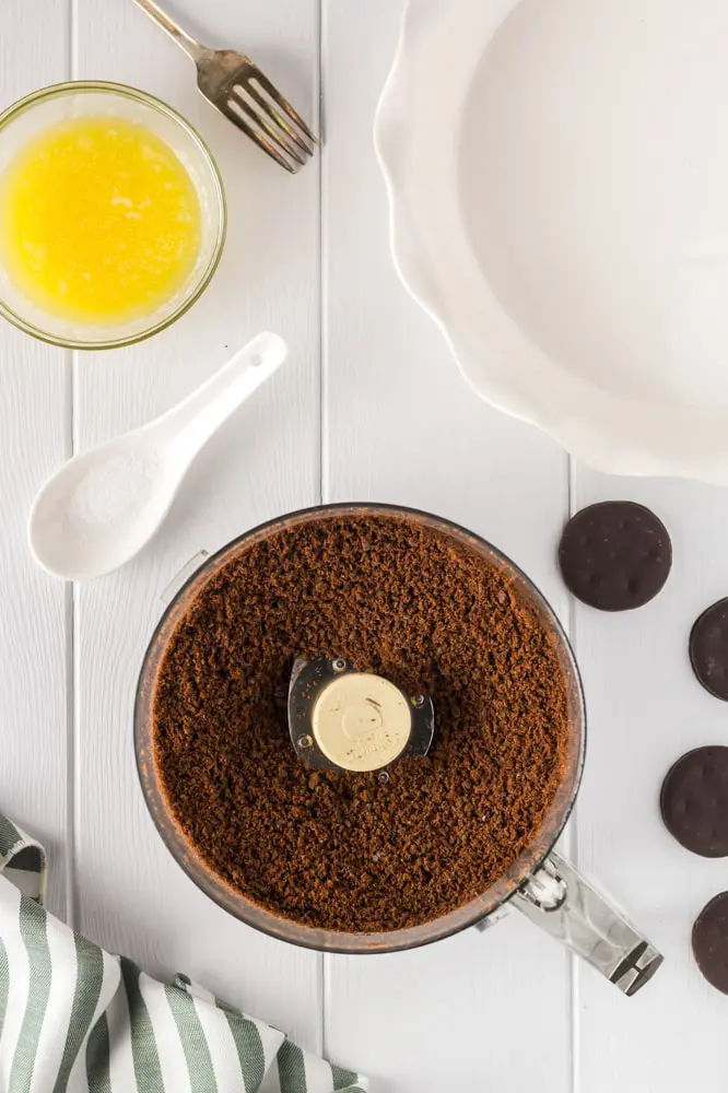 thin mint cookie crumbs in a food processor with a small bowl of melted butter and an empty white pie plate on the side