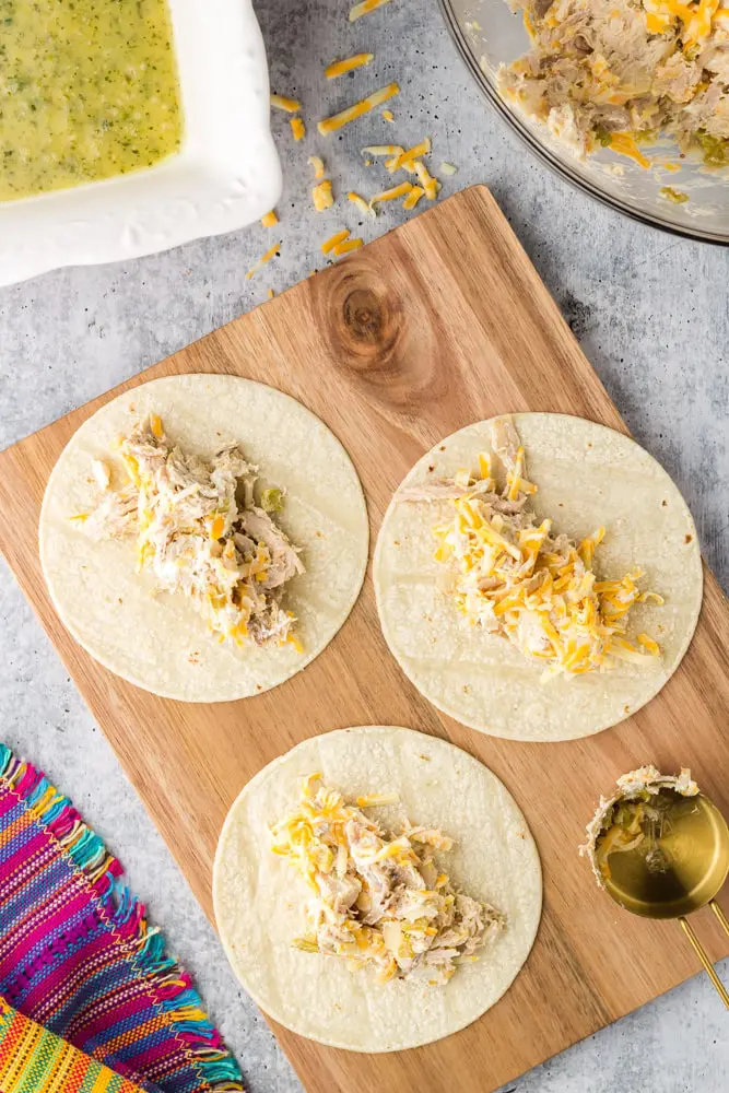 three white corn tortillas topped with chicken enchilada filling on a wood cutting board