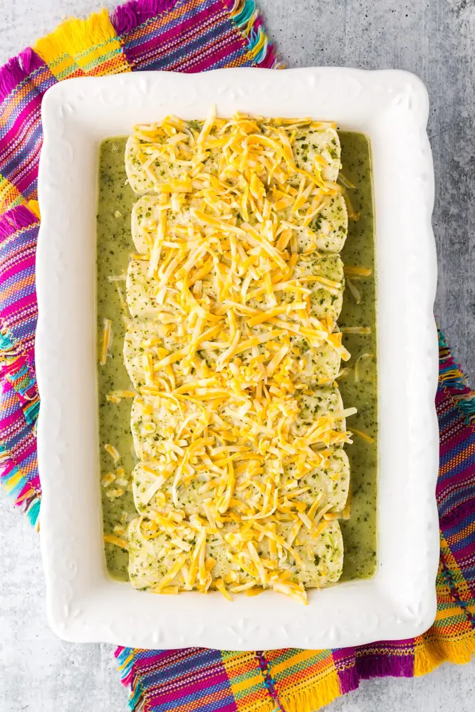 white baking dish filled with green chicken enchiladas topped with shredded cheese before being baked