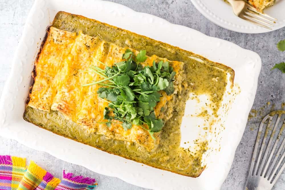 white baking dish filled with green chicken enchiladas topped with orange melted cheese and a mound of bright green cilantro with two enchiladas missing from the pan