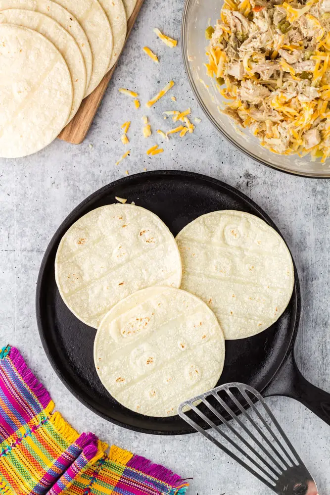 Three white corn tortillas on a black cast iron skillet with a silver spatula leaning on the pan