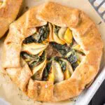 golden brown round sweet potato, apple and kale galette filled with orange slices of sweet potato, green torn kale leaves and white apple slices on a white baking sheet lined with brown parchment paper