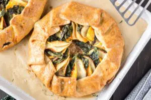 golden brown round sweet potato, apple and kale galette filled with orange slices of sweet potato, green torn kale leaves and white apple slices on a white baking sheet lined with brown parchment paper