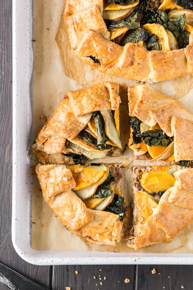 golden brown round sweet potato, apple and kale galette filled with orange slices of sweet potato, green torn kale leaves and white apple slices on a white baking sheet lined with brown parchment paper