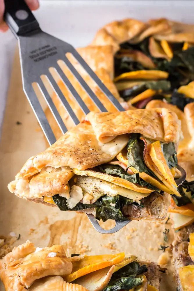 golden brown round sweet potato, apple and kale galette filled with orange slices of sweet potato, green torn kale leaves and white apple slices on a white baking sheet lined with brown parchment paper