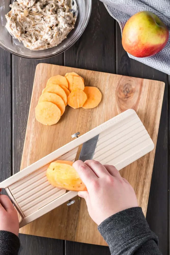 orange sweet potatoes being cut with a mandoline on a wooden cutting board