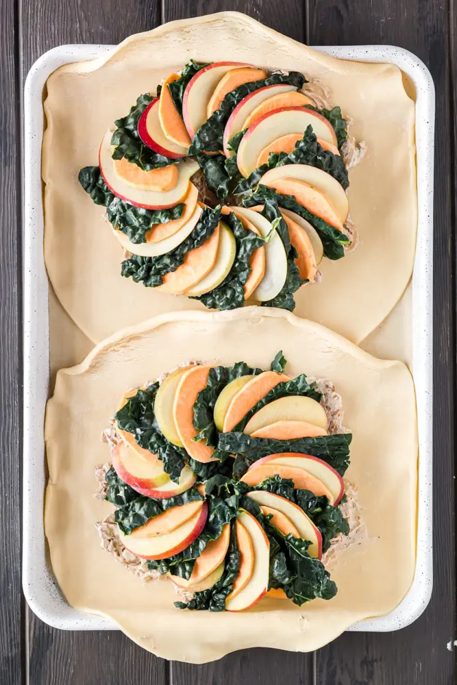 golden brown round sweet potato, apple and kale galette filled with orange slices of sweet potato, green torn kale leaves and white apple slices on a white baking sheet lined with brown parchment paper before folding edges of the pie crust over the vegetables