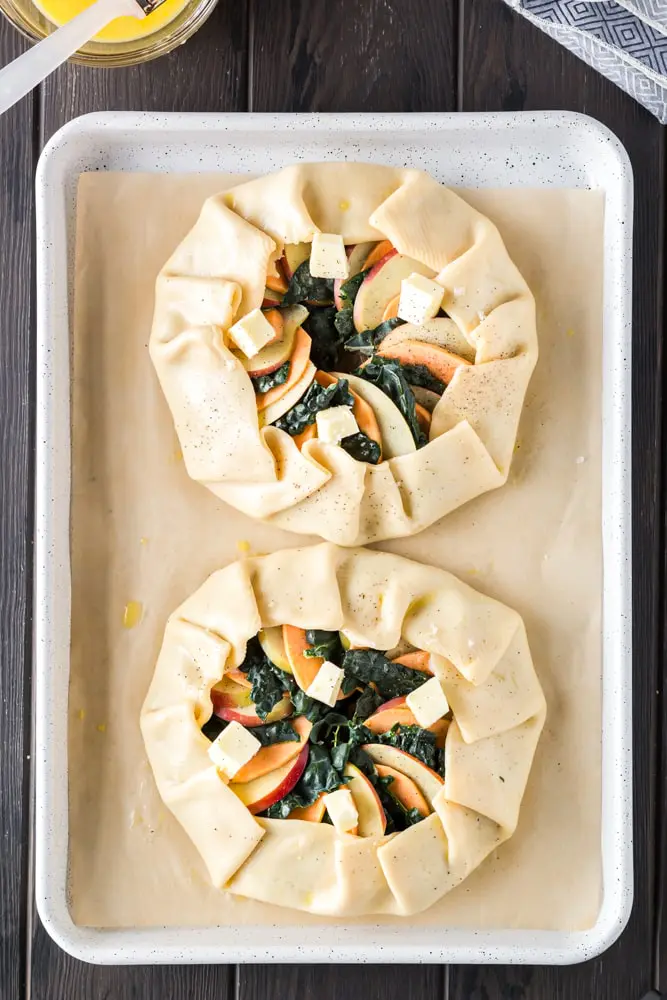 golden brown round sweet potato, apple and kale galette filled with orange slices of sweet potato, green torn kale leaves and white apple slices on a white baking sheet lined with brown parchment paper before baking