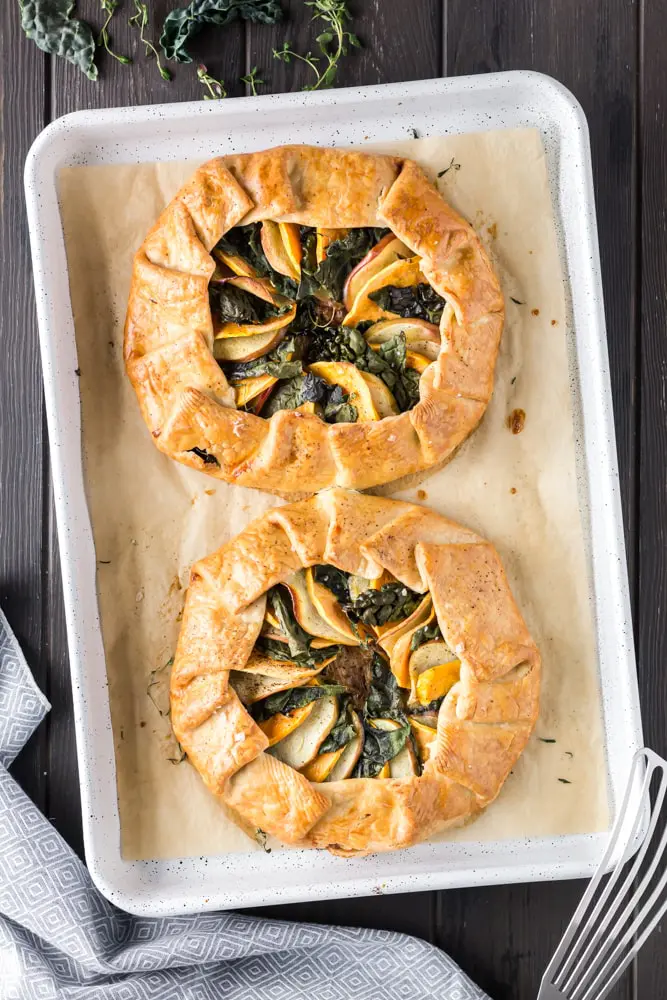 golden brown round sweet potato, apple and kale galette filled with orange slices of sweet potato, green torn kale leaves and white apple slices on a white baking sheet lined with brown parchment paper