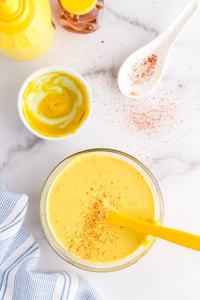 Bright yellow honey mustard dressing in a glass bowl being stirred by a yellow spatula.