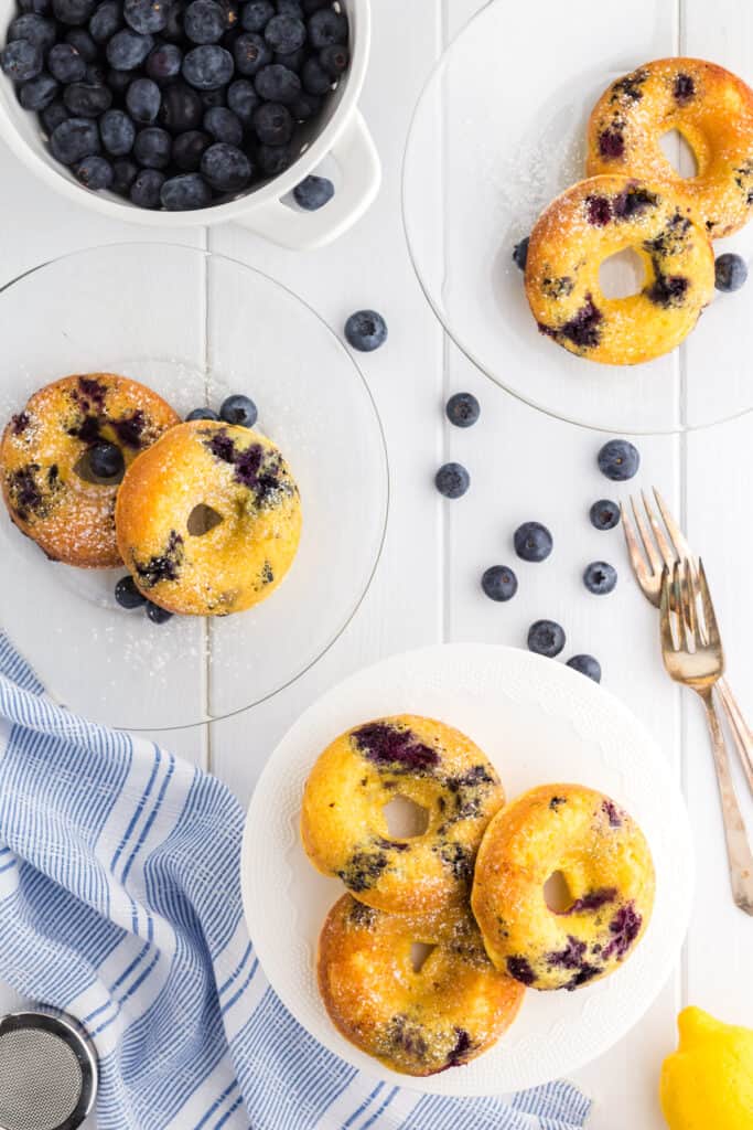 Two servings of blueberry cake donuts on glass plates from a white pedestal of donuts.