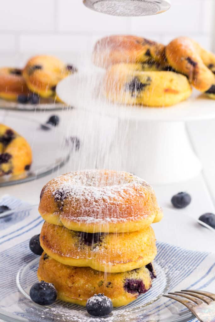 3 Baked Blueberry Cake Donuts in a stack on a clear glass plate being dusted with powdered sugar.