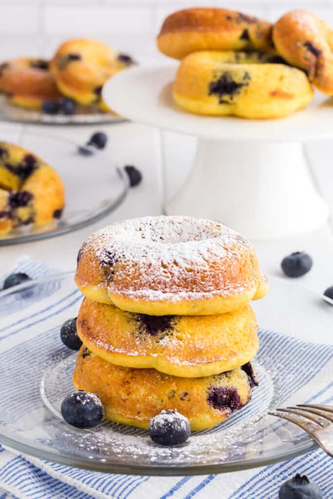 3 Baked Blueberry Cake Donuts in a stack on a clear glass plate dusted with powdered sugar.