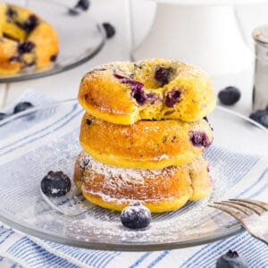 3 Baked Blueberry Cake Donuts in a stack on a clear glass plate dusted with powdered sugar. There is bite taken out of the donut on top.