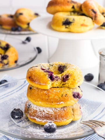 3 Baked Blueberry Cake Donuts in a stack on a clear glass plate dusted with powdered sugar. There is bite taken out of the donut on top.