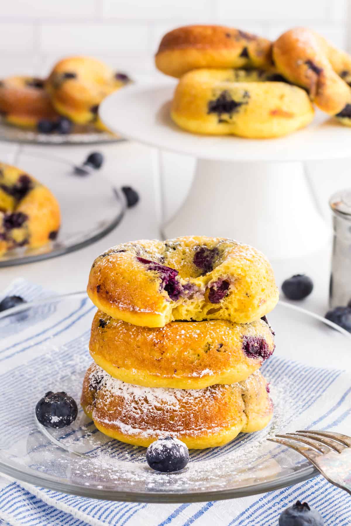 3 Baked Blueberry Cake Donuts in a stack on a clear glass plate dusted with powdered sugar. There is bite taken out of the donut on top.