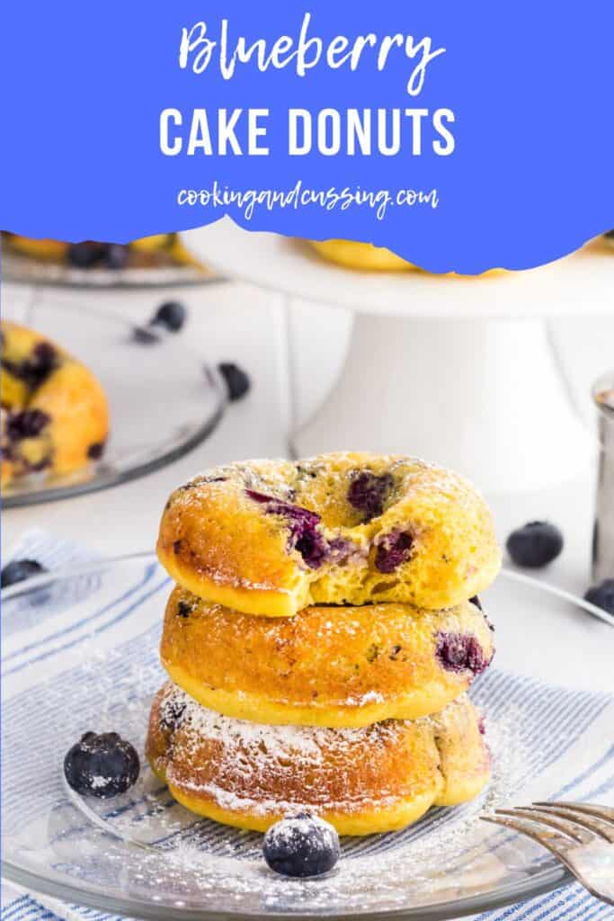 3 Baked Blueberry Cake Donuts in a stack on a clear glass plate dusted with powdered sugar. There is bite taken out of the donut on top.