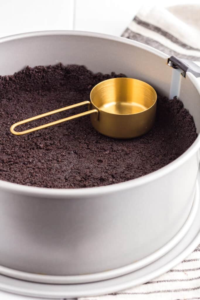 The oreo cookie crust being pressed into the spring form pan with a measuring cup to create a clean edge.