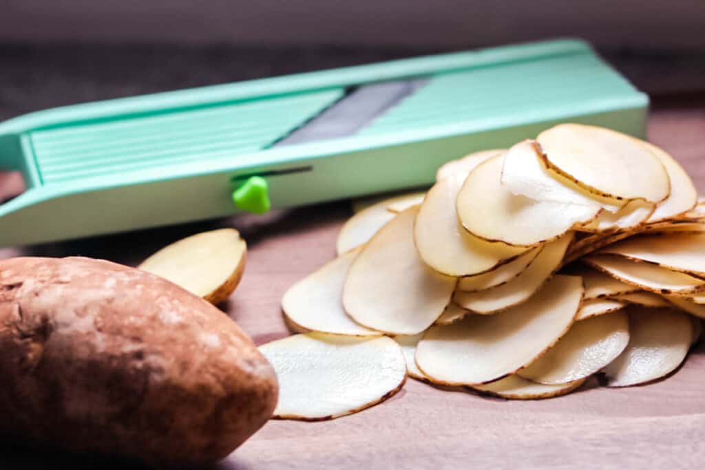 Thinly sliced potatoes on a cutting board with a mandoline slicer.