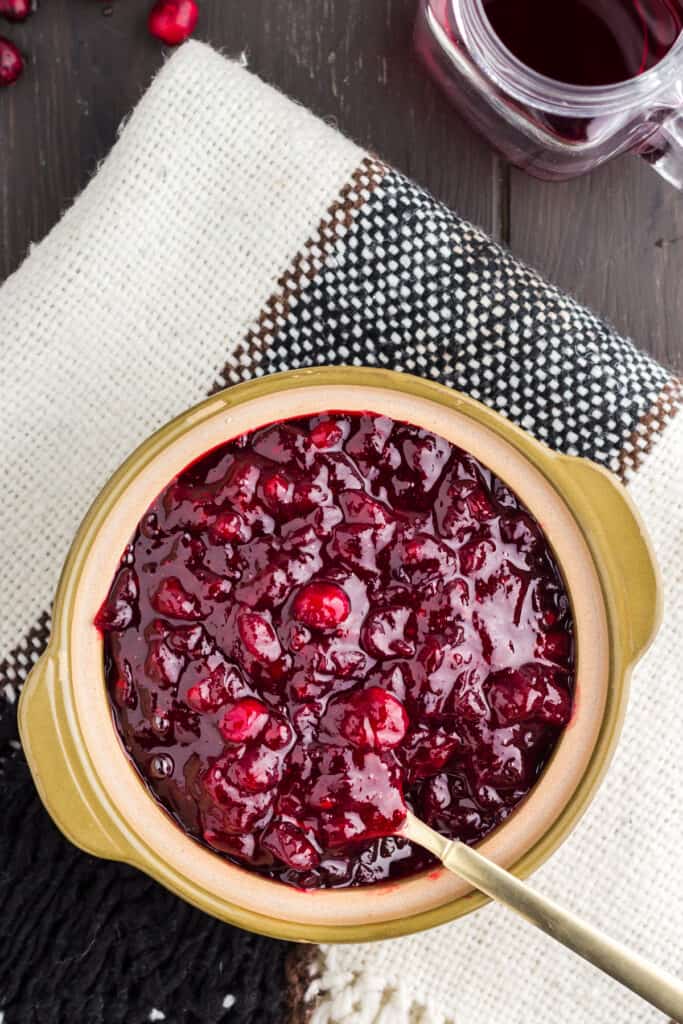 Homemade cranberry sauce with grand marnier in an olive green bowl with a gold spoon on top of black and white tablecloth.