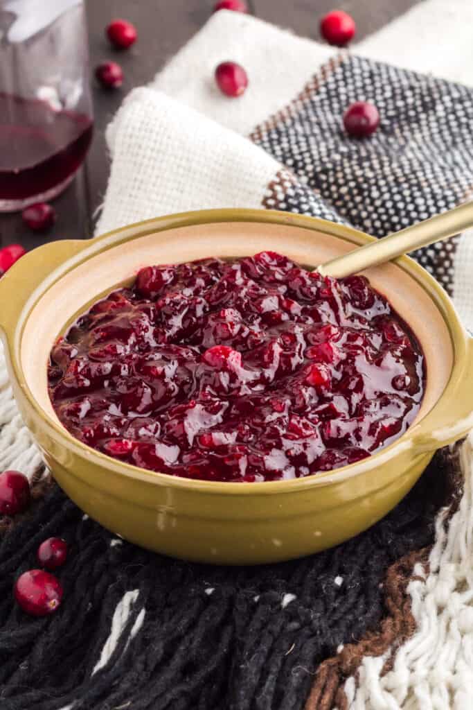 Homemade cranberry sauce with grand marnier in an olive green bowl with a gold spoon on top of black and white tablecloth.