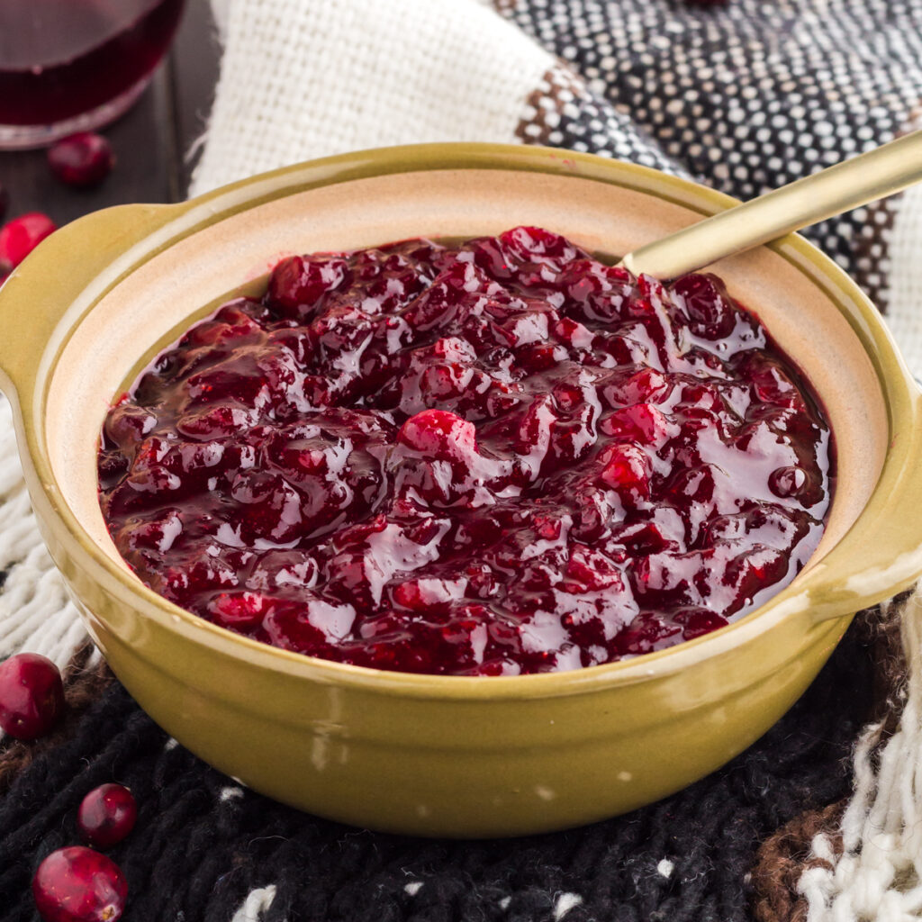 Homemade cranberry sauce with grand marnier in an olive green bowl with a gold spoon on top of black and white tablecloth.