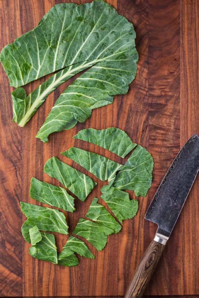 Two collard green leaves on a dark wood cutting board. One has the stem removed and the other is cut into pieces.