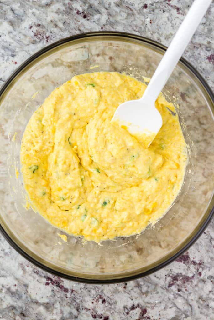 Cornbread batter being mixed in a glass bowl with a white spatula.