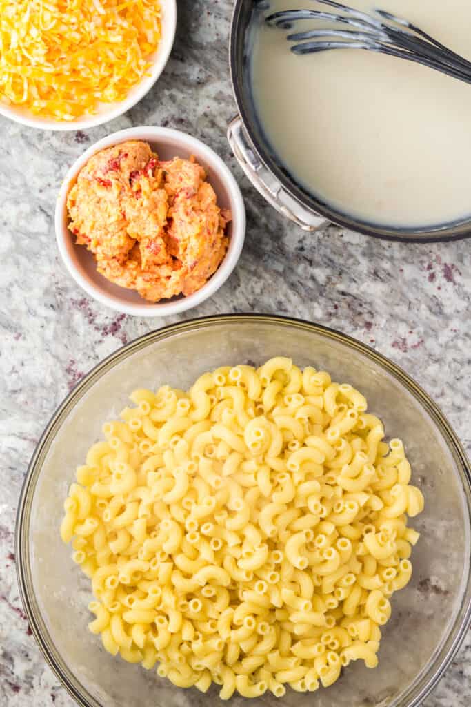 Cooked noodles in a large glass bowl with white sauce, pimento cheese and shredded cheese on the side.